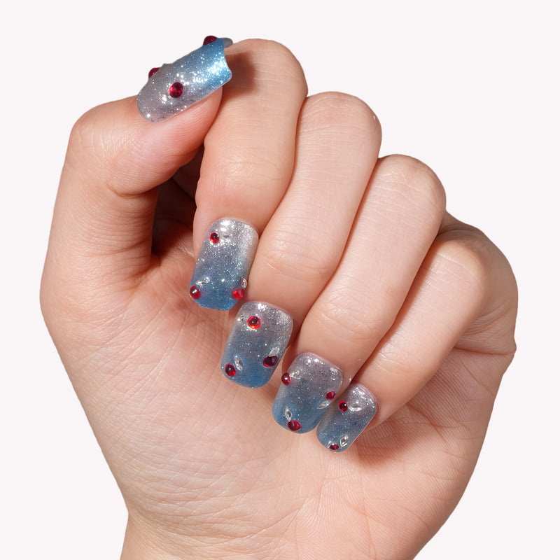 Hand model wearing Frosted Cherry press-on nails in silver and blue with red gem accents on a white background