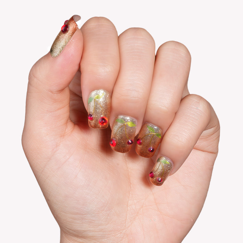Hand model wearing Frosted Cherry press-on nails in gold and green glitter with red gem accents on a white background
