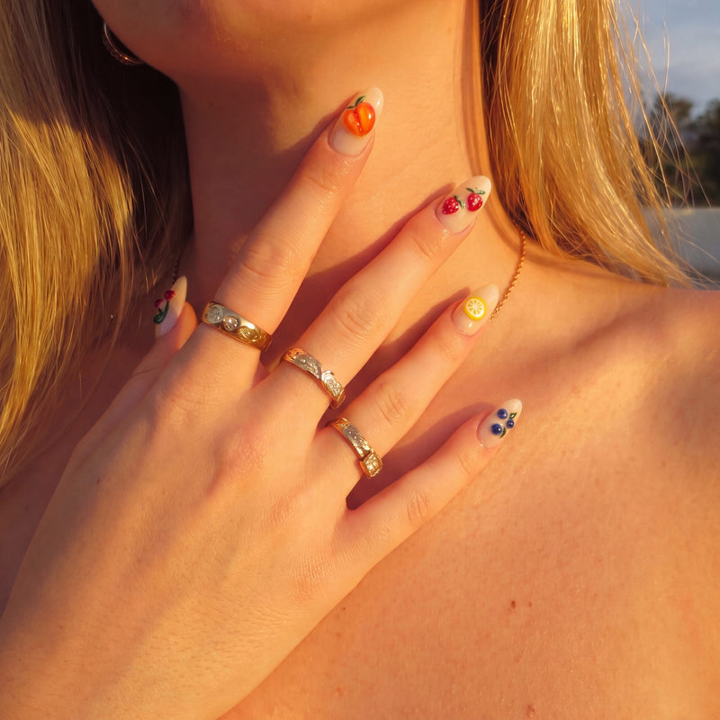 Close-up of Fruit Crush cream nails with 3D peach, cherries, lemon, and blueberries in sunset light with gold rings
