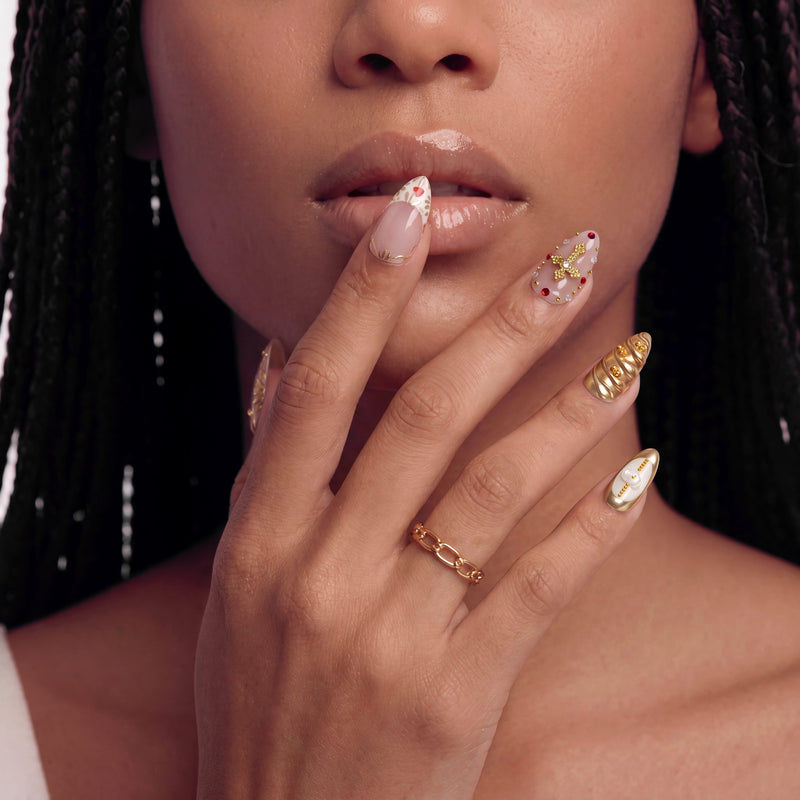 Close-up of a hand model wearing blush pink and gold jeweled press-on nails near the lips.