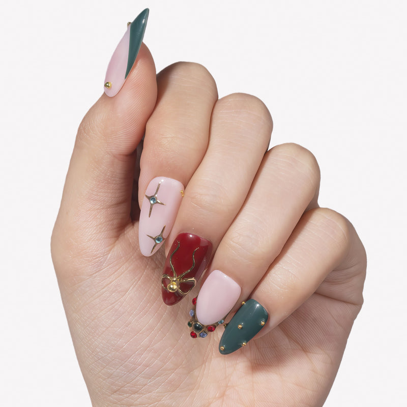 Hand model wearing Gilded Noel press-on nails with burgundy, deep teal, nude pink, gold bow, starburst, and jewel accents