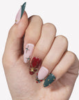 Hand model wearing Gilded Noel press-on nails with burgundy, deep teal, nude pink, gold bow, starburst, and jewel accents