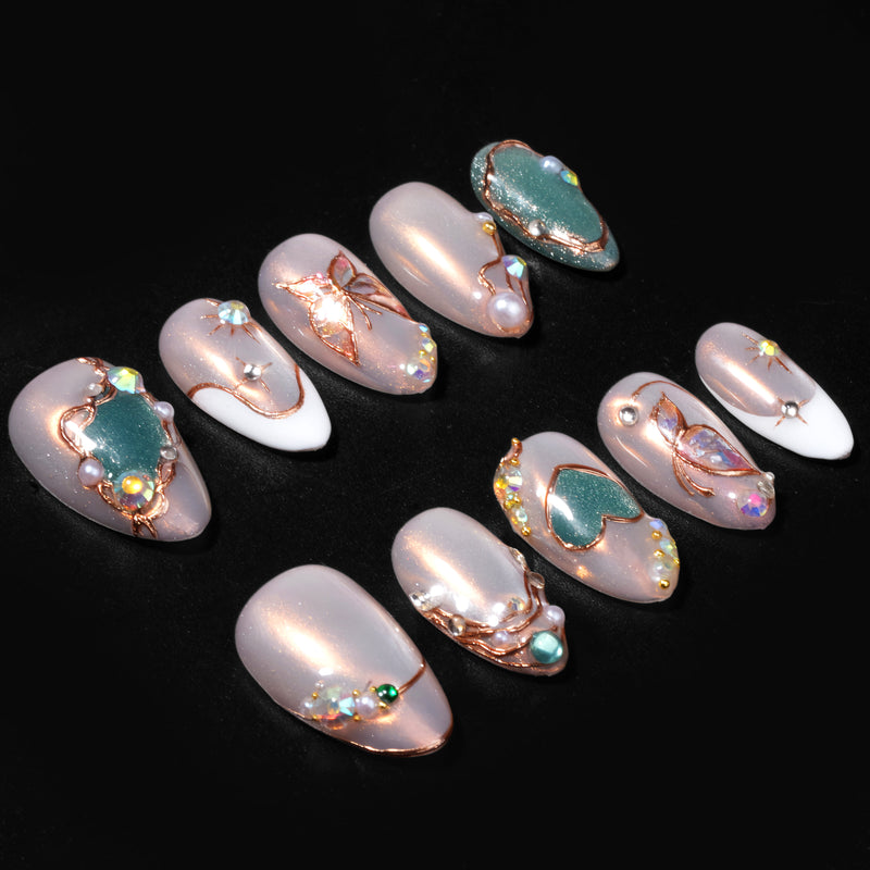 A black background flat lay of champagne shimmer press-on nails with olive green heart accents, pearls, and rhinestones