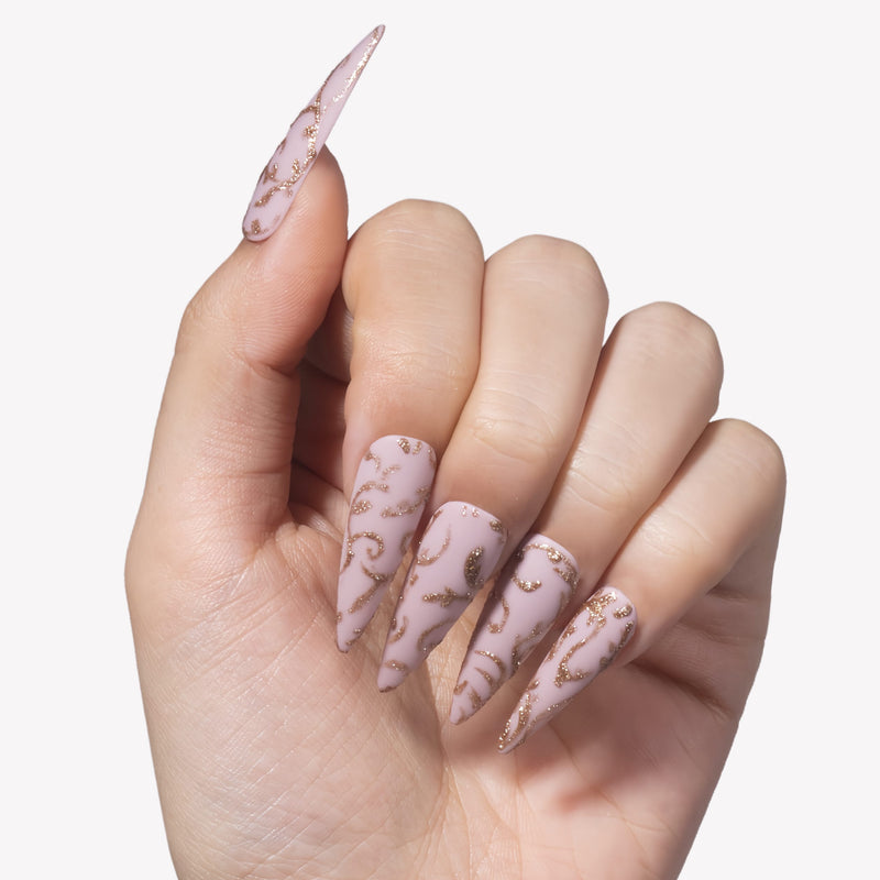 Close-up hand model wearing Gilded Petals stiletto press-on nails with soft pink bases and raised gold vine patterns