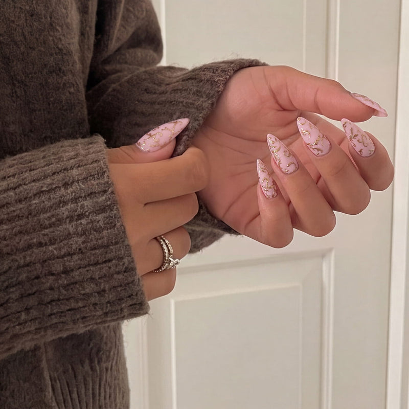 Hand model wearing Gilded Petals press-on nails with soft pink bases and raised gold vine patterns beside a cozy sweater sleeve