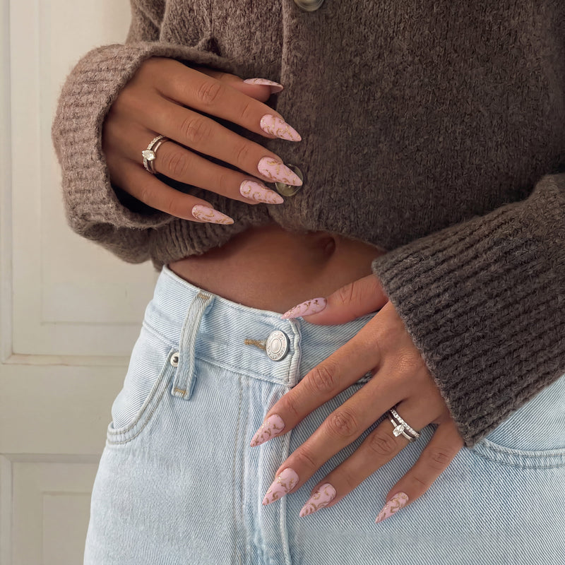 Lifestyle hand model wearing Gilded Petals press-on nails with pink bases and raised gold vine designs with a sweater and jeans