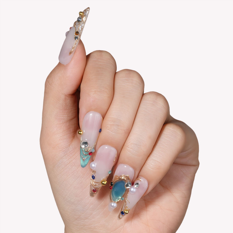Close-up of a hand model wearing Gilded Relic Blue press-on nails with blue gems, pearls, and gold accents