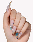 Close-up of a hand model wearing Gilded Relic Blue press-on nails with blue gems, pearls, and gold accents