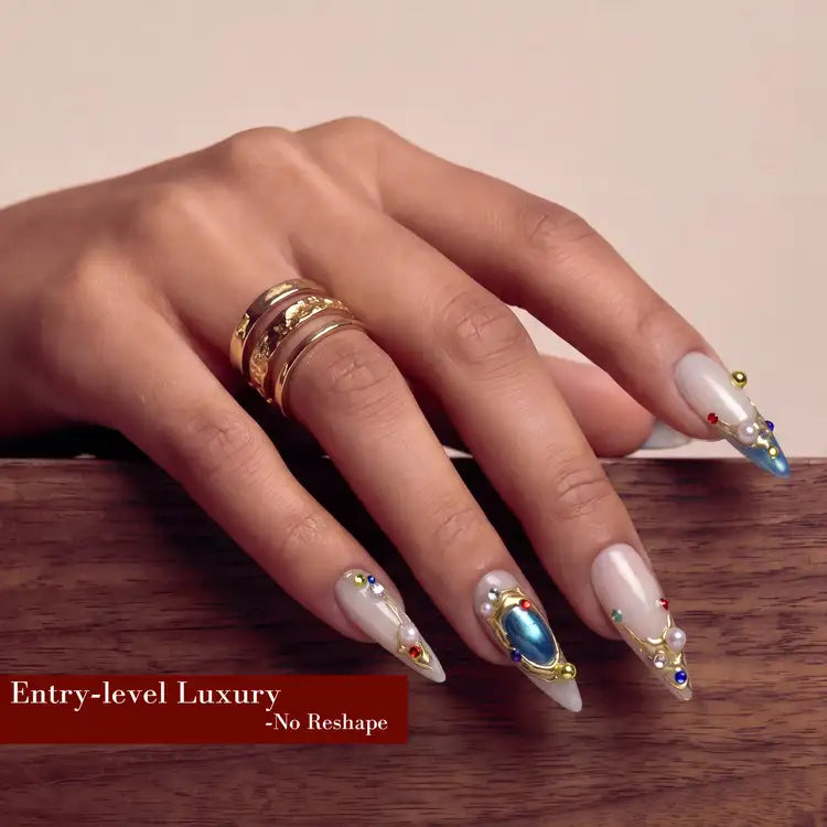 Hand model wearing long nude press-on nails with blue gemstone accents and gold rings on a wooden surface.