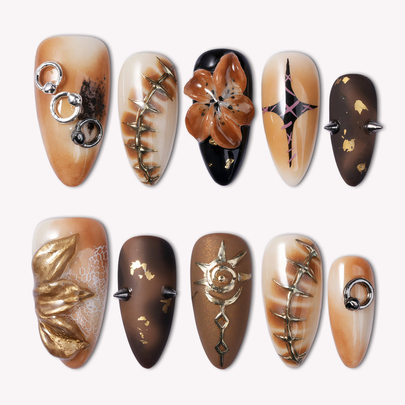 A flat lay of long almond press-on nails in warm brown and gold tones with metallic rings, spikes, and 3D floral accents.