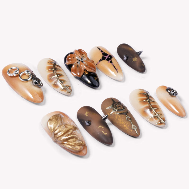 A flat lay of brown and gold press-on nails with metallic rings, spikes, florals, and abstract accents on a white background.