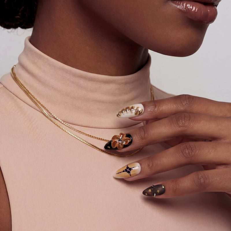 A hand model wearing brown and gold press-on nails poses against a beige top and gold necklace.