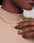 A hand model wearing brown and gold press-on nails poses against a beige top and gold necklace.