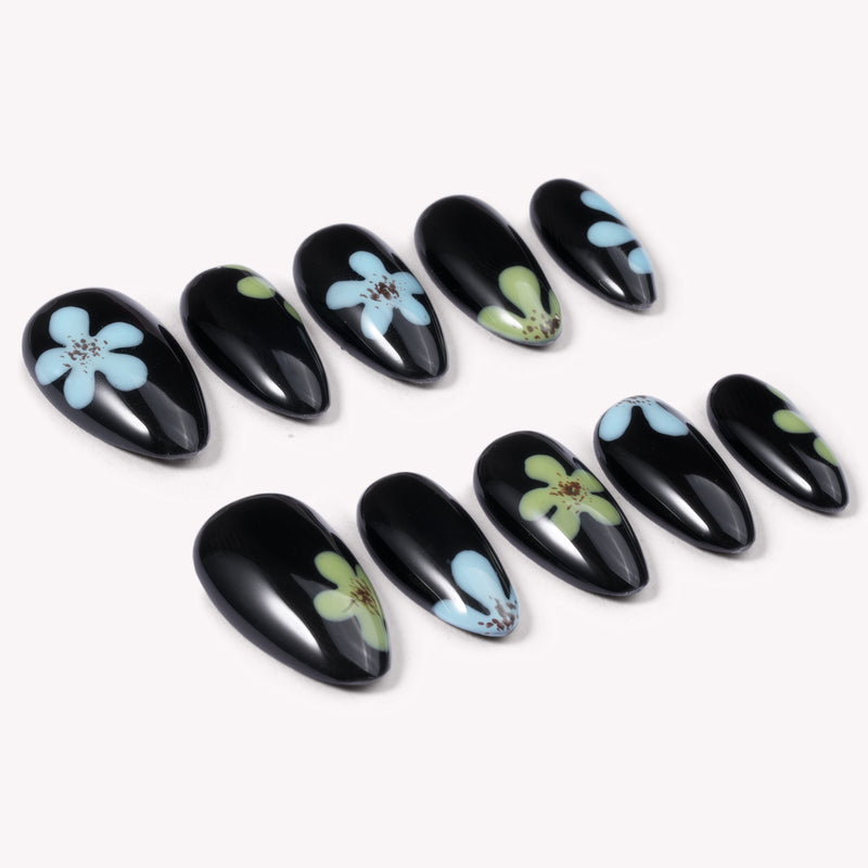 A flat lay of black press-on nails with blue and green flower accents arranged in two rows on a white background.