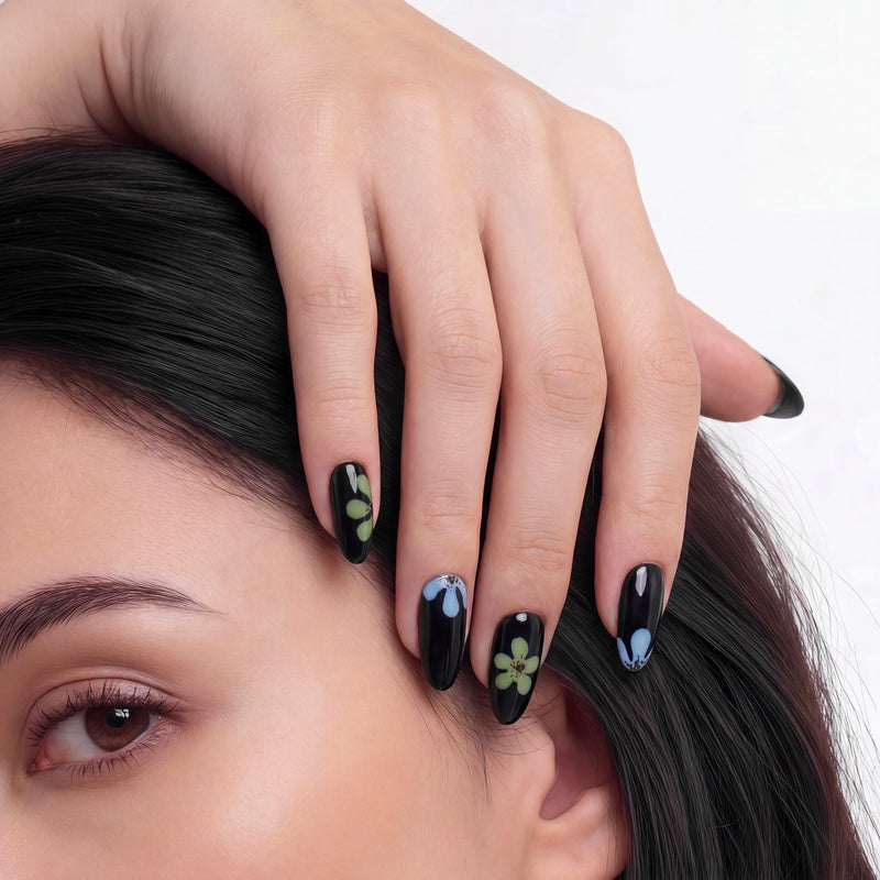 A hand wearing glossy black press-on nails with blue and green flower accents poses over a woman's face.