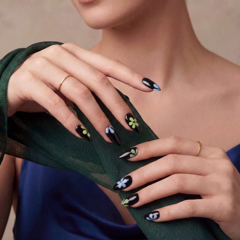 A model wearing glossy black press-on nails with blue and green flower accents poses with a dark green scarf.