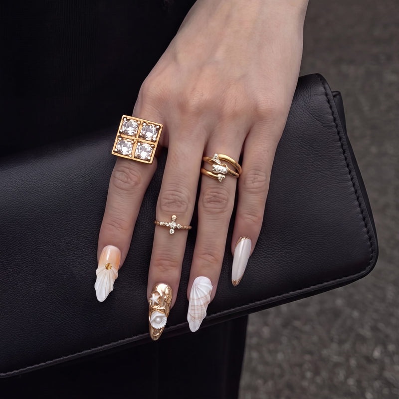 Hand close-up wearing Golden Shell white and gold seashell nails with gold rings on a black clutch