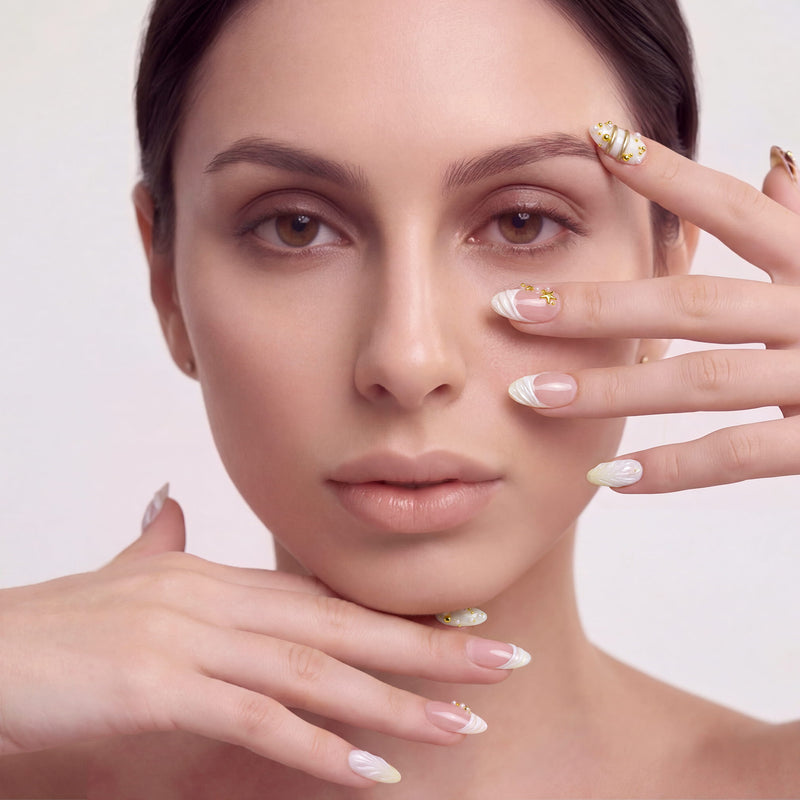 Hand model wearing Golden Shore Ritual handmade press-on nails with pearl white, blush pink, and gold accents on a botanical background.