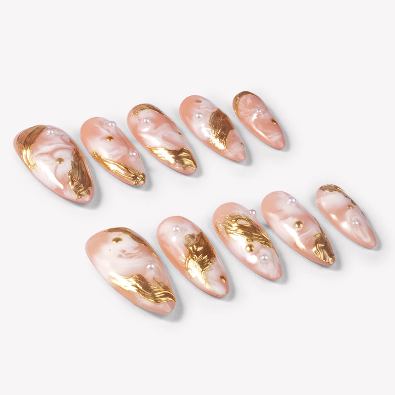Angled flat lay of Golden Veil peach marble press-on nails with metallic gold foil, pearls, and gold studs