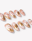 Angled flat lay of Golden Veil peach marble press-on nails with metallic gold foil, pearls, and gold studs
