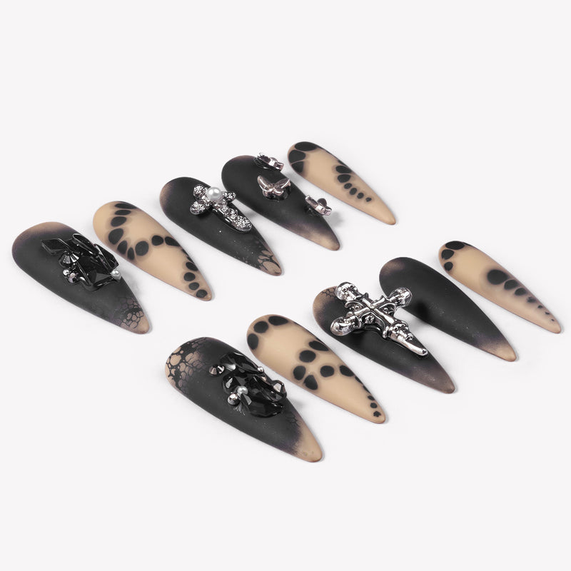 Angled flat lay of Gothic Elegance press-on nails with matte black and nude dot designs, silver cross charms, black rhinestones, lace effects, and butterflies