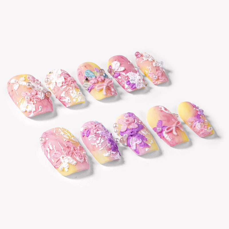 Angled flat lay of Impressionist Pointe press-on nails with pink yellow gradients, 3D flowers, pearls, and bow accents