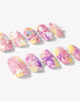 Angled flat lay of Impressionist Pointe press-on nails with pink yellow gradients, 3D flowers, pearls, and bow accents
