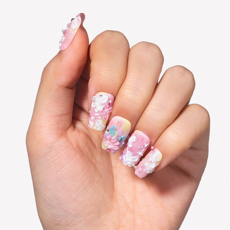 Hand model wearing Impressionist Pointe press-on nails with pink yellow gradients, 3D flowers, pearls, and bow accents