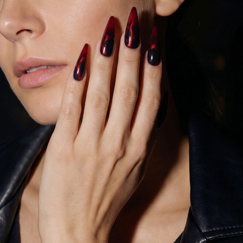 Side face hand model wearing Inferno Veins stiletto press-on nails with glossy deep red tips, matte black sections, and vein-like accents