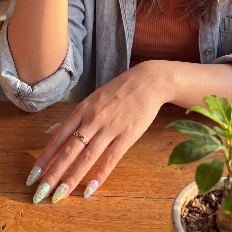 Hand model wearing Iridescent Petals pastel floral press-on nails beside a green plant on a wooden table
