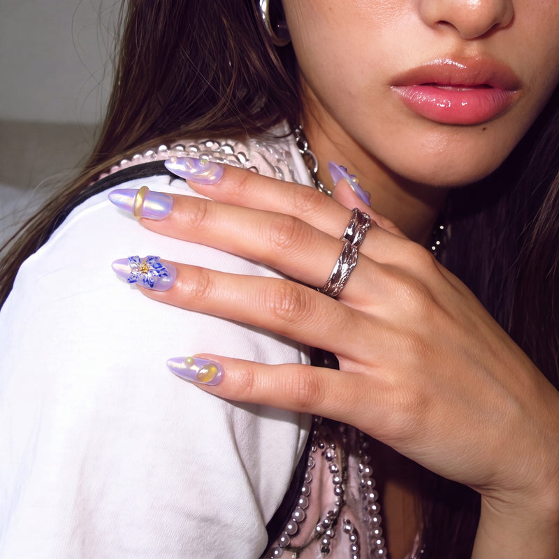 Fashion close-up of a model wearing Iris Bloom iridescent purple floral press-on nails with silver rings