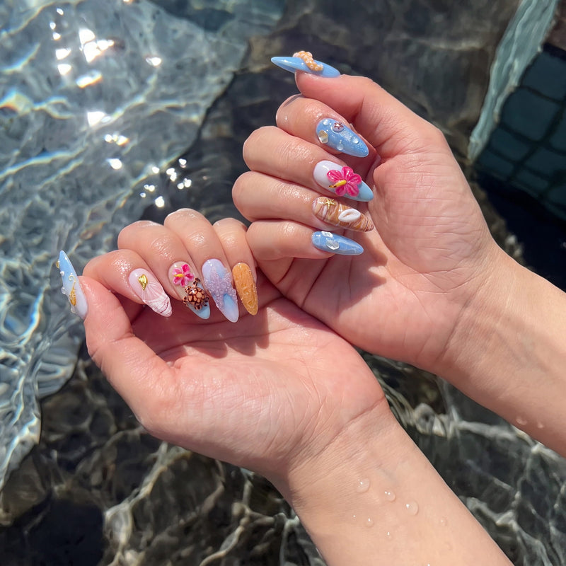 Both hands wearing Island Echo blue tropical press-on nails with pink flowers, shells and turtle charms beside pool water