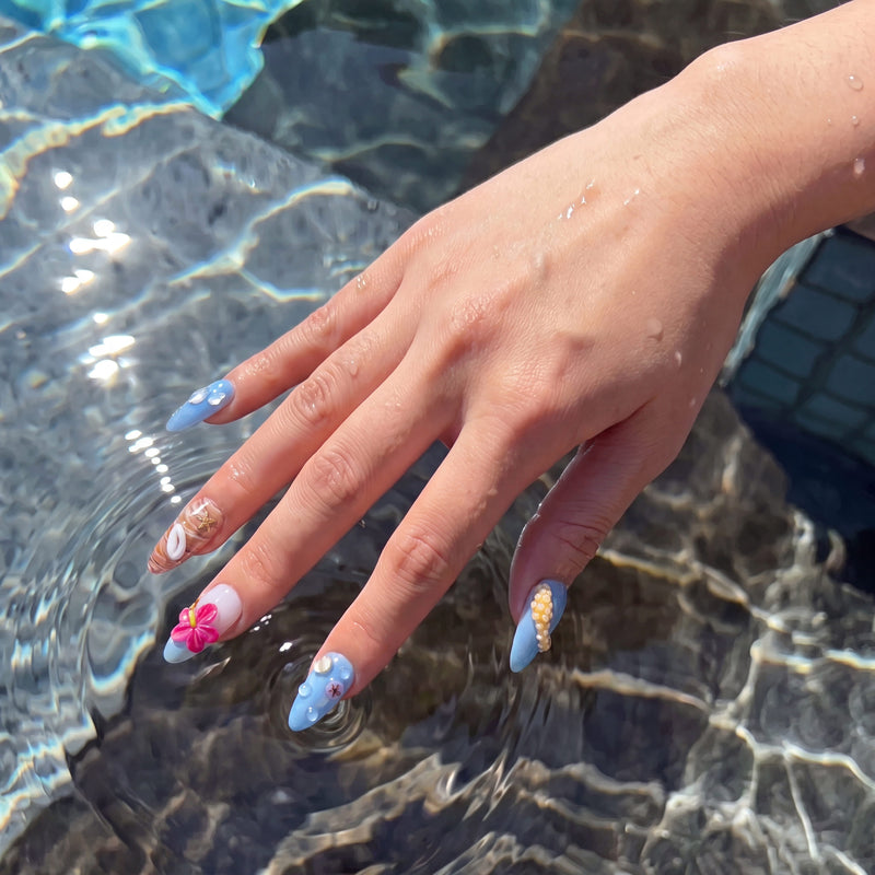 Wet hand wearing Island Echo blue tropical flower, shell and starfish press-on nails over rippling pool water
