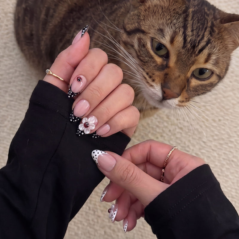 Two-hand model near a cat wearing Ladybug Garden press-on nails with black and white polka dot French tips, ladybug accents, and a white flower charm