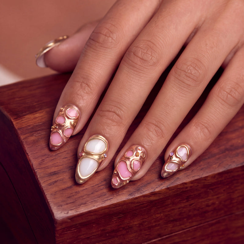 Hand model displaying Liquid Gold pink and white press-on nails with rose gold frames on a wooden box