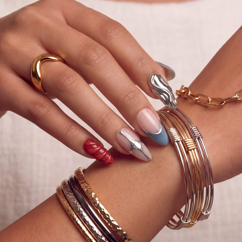 Close-up hand model wearing Liquid Mercury press-on nails with silver chrome texture, blue gray tips, red accent nail, and stacked bracelets
