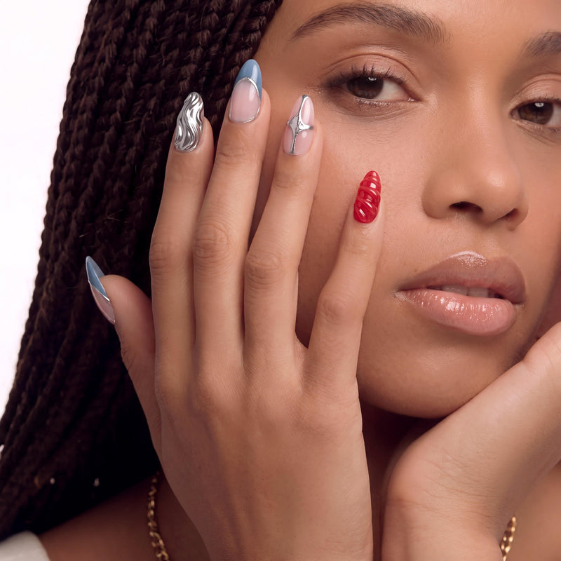 Face close-up hand model wearing Liquid Mercury press-on nails with silver chrome swirls, blue gray tips, and a red textured accent nail