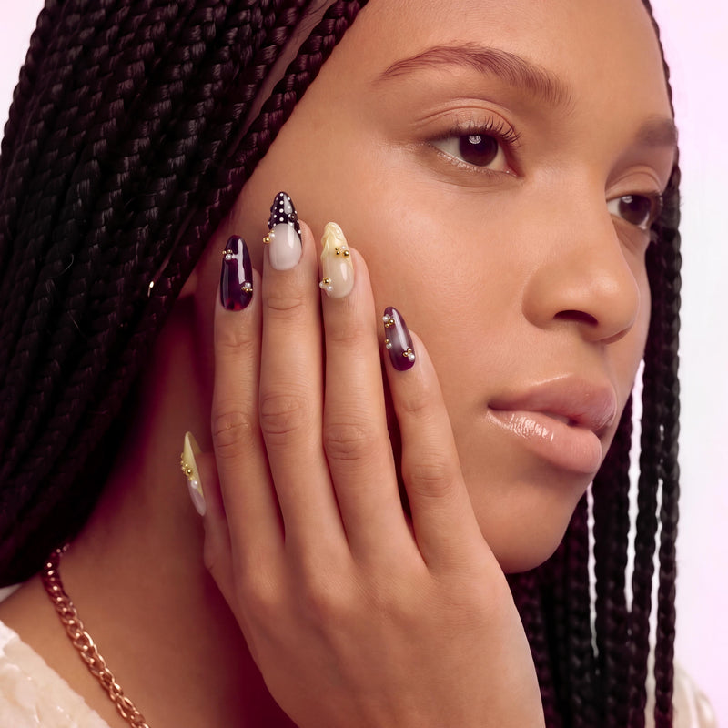 Face close-up hand model wearing Lunar Velvet press-on nails with pale yellow tips, burgundy cat eye polish, black dotted lattice, gold beads, and pearls