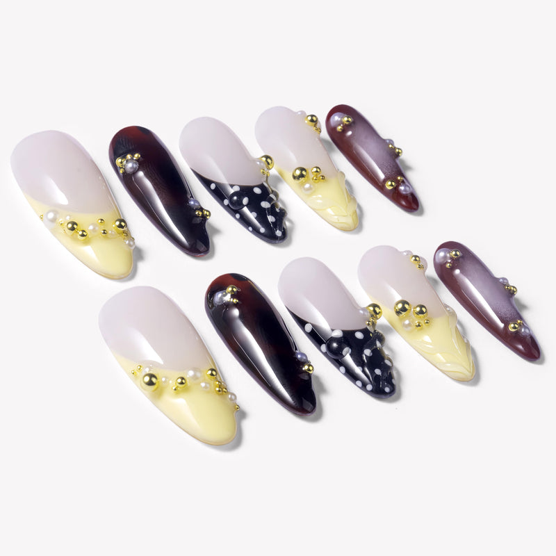 Flat lay of Lunar Velvet press-on nails with pale yellow tips, black dotted lattice designs, burgundy cat eye polish, pearls, and gold bead accents