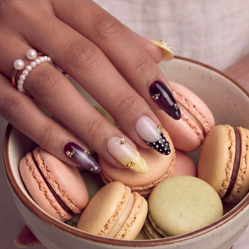 Hand model holding macarons while wearing Lunar Velvet press-on nails with burgundy cat eye polish, pale yellow tips, black dotted lattice, pearls, and gold beads