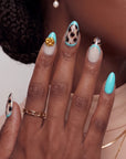Hand model wearing turquoise and cream almond press-on nails with black abstract spots and a gold flower accent