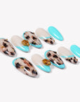 Two rows of turquoise and cream almond press-on nails with black abstract spots and gold flower accents arranged on a white background