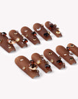 Angled flat lay of Luxurious Treasure press-on nails in a warm brown square style with gold, amber, and clear rhinestone gems