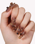 Hand model wearing Luxurious Treasure press-on nails in a warm brown square style with gold, amber, and clear rhinestone gems