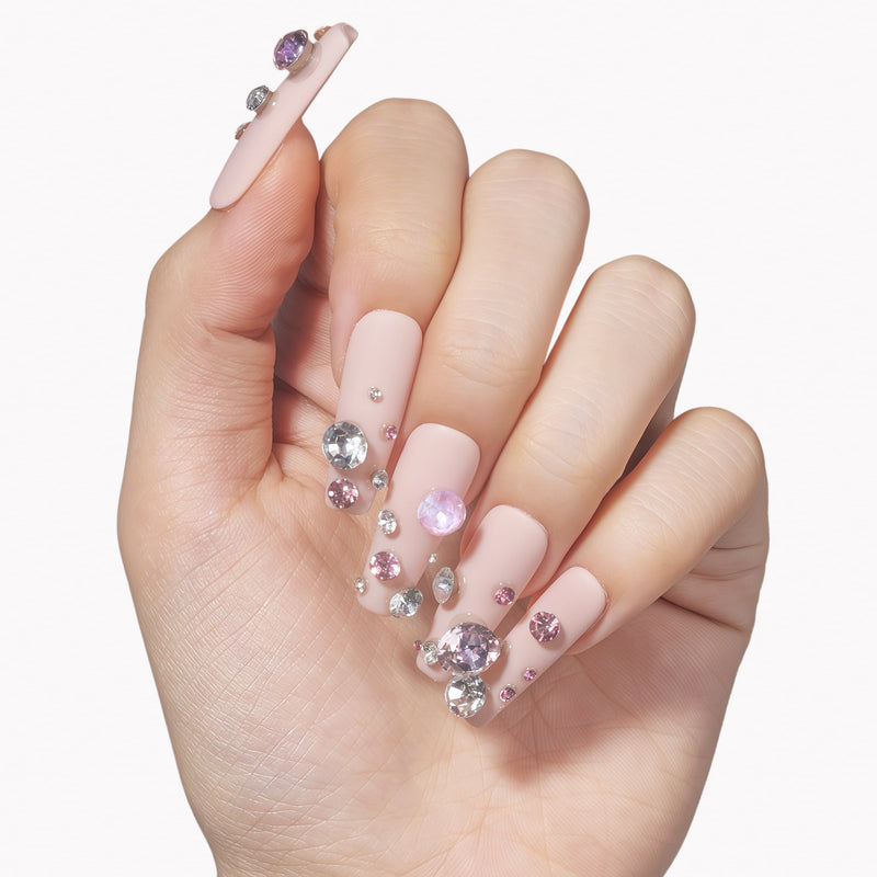 Hand model wearing Luxurious Treasure press-on nails with nude pink square bases and large pink and clear rhinestone gems on a white background