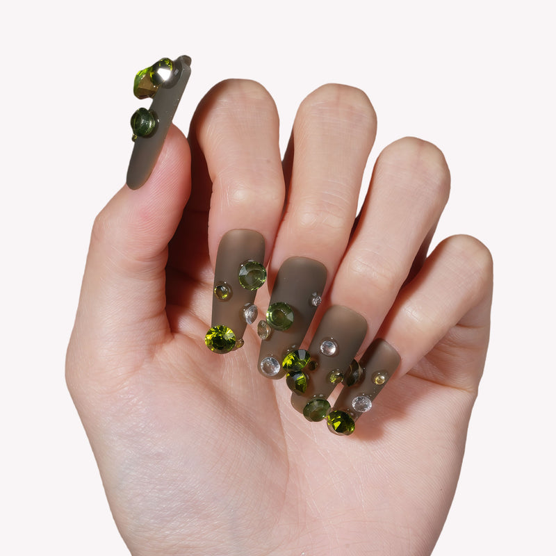 Hand model wearing Luxurious Treasure press-on nails in an olive green square style with large green and clear rhinestone gems
