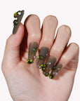 Hand model wearing Luxurious Treasure press-on nails in an olive green square style with large green and clear rhinestone gems