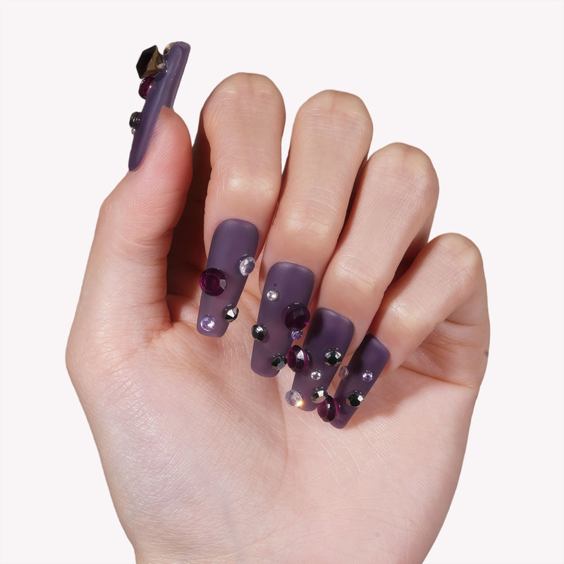 Hand model wearing Luxurious Treasure press-on nails in a muted purple square style with dark purple, black, and clear rhinestone gems