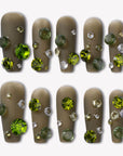 Flat lay of Luxurious Treasure press-on nails in an olive green square style with large green, yellow, and clear rhinestone gems