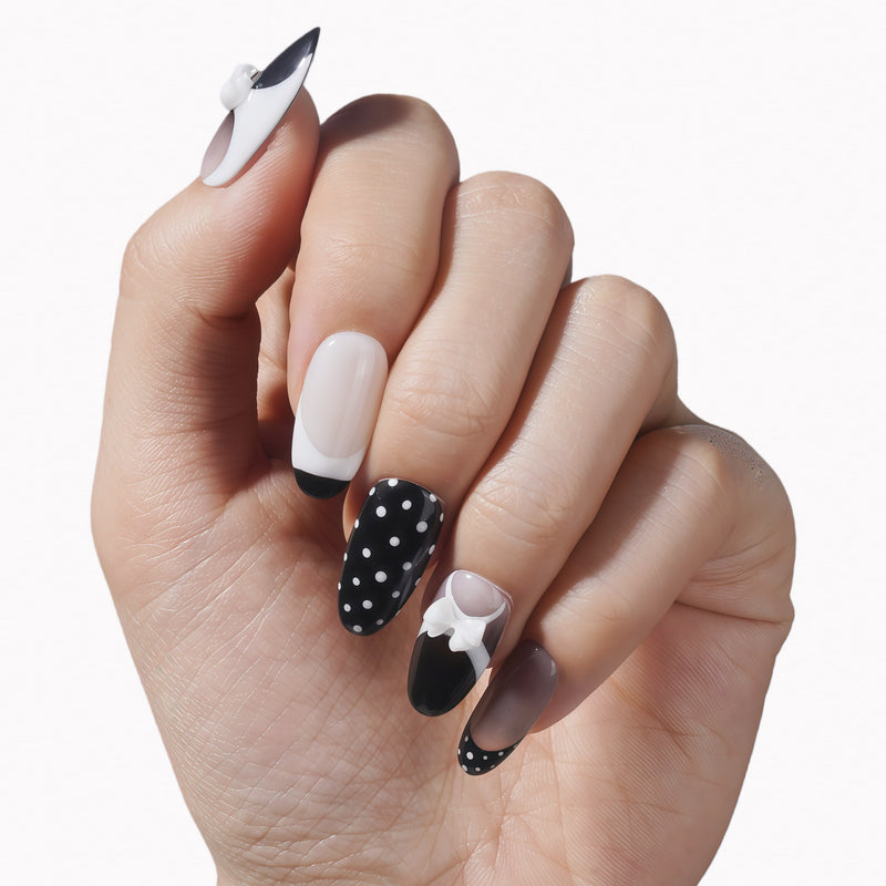 Hand model wearing black and white press-on nails with polka dots and white bow accents